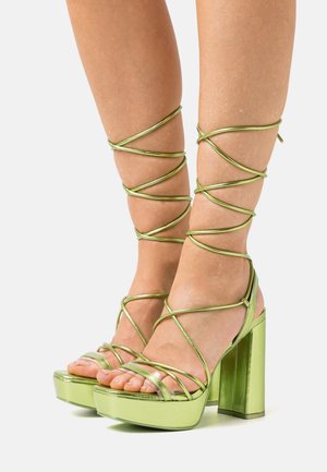 Sandalias de tacón alto metálicas verdes con una plataforma gruesa, con tiras entrelazadas alrededor del tobillo y diseño de punta descubierta.