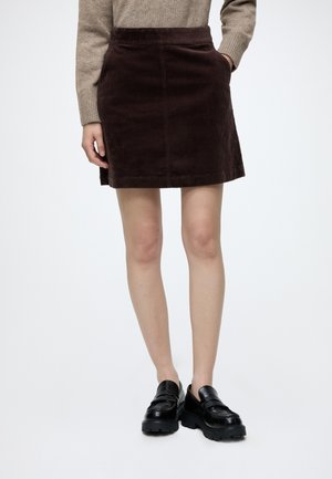 Person, die einen braunen Cord-Minirock, einen beigen Pullover und schwarze klobige Loafers trägt und mit einer Hand in der Tasche steht.