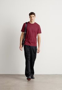 Jonge man die vooruit loopt met een bordeauxrood T-shirt, zwarte broek, zwarte schoenen en een zwarte schoudertas op een effen achtergrond.