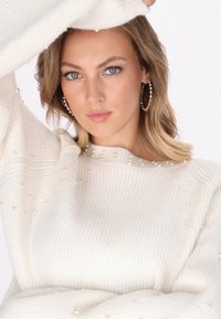 Pulover alb cu dungi, decorat cu mici accente de perlă, având un decolteu rotund și o croială lejeră. Textură netedă, material moale și mâneci lungi.