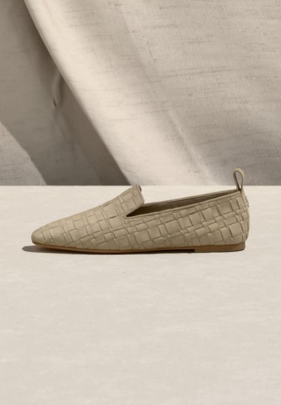 Πλεκτό μπεζ επίπεδο παπούτσι χωρίς κορδόνια με μυτερή μύτη και θηλιά στο πίσω μέρος, πάνω σε ουδέτερο υφασμάτινο φόντο.