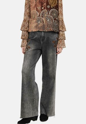 Persona que lleva vaqueros de pierna ancha negros desgastados con bordados florales cerca de los bolsillos y una blusa marrón estampada de manga larga con puños con volantes.