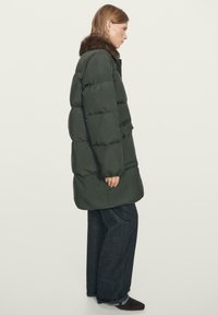 Woman wearing a dark green puffer coat with fur collar, dark wide-leg jeans, and dark shoes, standing sideways against a plain background.
