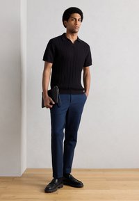 Μαύρο πλεκτό πόλο με κοντά μανίκια και ριμπ υφή, συνδυασμένο με ναυτικό μπλε κωνικά παντελόνια και μαύρα μοκασίνια, κρατώντας μαύρο τσαντάκι clutch.