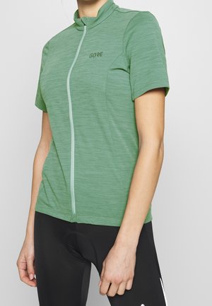 Cyklist iført en tætsiddende grøn kortærmet cykeltrøje med lynlås og sorte cykelshorts med polstring og hvidt logo på låret.