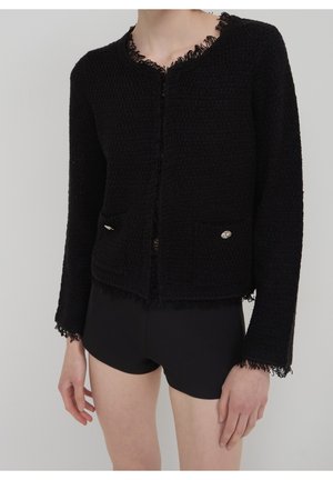 Femme portant une veste noire texturée avec des bords en dentelle et des boutons décoratifs, associée à un short noir ajusté sur un fond uni.