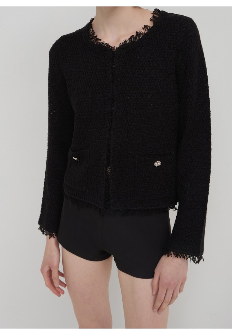 Femme portant une veste noire texturée avec des bords en dentelle et des boutons décoratifs, associée à un short noir ajusté sur un fond uni.
