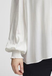 Weiße Bluse mit vertikalen Streifen, puffartigen Ärmeln und gesmokten Bündchen. Der Stoff wirkt leicht und strukturiert, mit einer fließenden Silhouette.