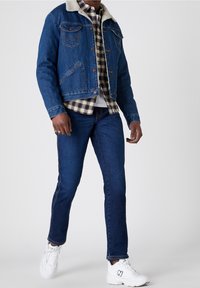 Denim jack met een shearling kraag, blauwe kleur, met twee borstzakken en knoopsluitingen, gedragen over een geruit shirt en donkere jeans.