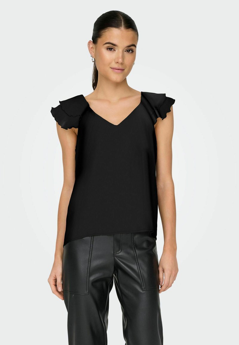 Jeune femme portant un haut noir sans manches à col en V avec des épaules volantées et un pantalon noir taille haute en simili cuir, sur un fond uni.