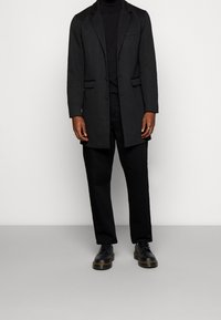 Cappotto nero su misura con rever a punta, due tasche frontali, abbinato a un dolcevita nero, pantaloni e scarpe stringate nere.