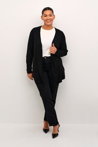 Cárdigan negro abierto sobre una blusa blanca, combinado con jeans negros y tacones puntiagudos negros. Textura suave y corte relajado, con mangas largas.
