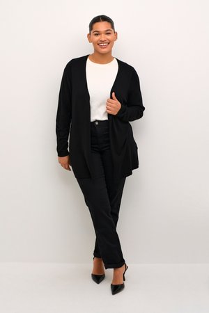 Cardigan noir ouvert sur un haut blanc, associé à un jean noir et des escarpins noirs à bout pointu. Texture lisse et coupe décontractée, avec des manches longues.