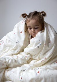 Bawełniany koc w kolorze kremowym, z kolorowymi ilustracjami zabawek i balonów. Miękka tekstura, owinięty wokół dziecka.