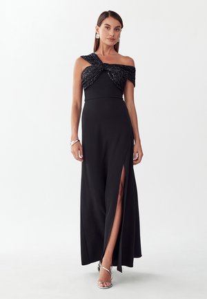 Frau trägt ein schwarzes schulterfreies Kleid mit Pailletten-Twist-Corsage und einem Oberschenkelschlitz, kombiniert mit silbernen Riemchenabsätzen und Accessoires.