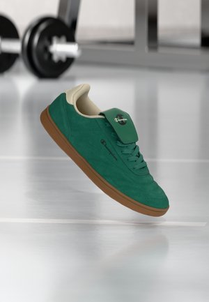 Green suede sneaker with brown sole and Champion logo, suspended mid-air in a gym with weights in soft focus background.