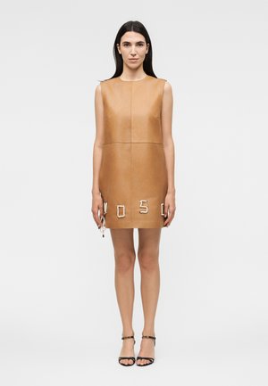 Hosszú sötét hajú nő, aki ujjatlan, világosbarna bőr miniruhát visel, amelynek szegélye közelében kötélből készült betűk vannak, valamint fekete pántos magassarkúkat.