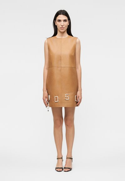 Femme aux longs cheveux foncés portant une mini-robe en cuir marron clair sans manches avec des lettres en corde près de l’ourlet et des talons noirs à brides.