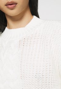 Weißer gestrickter Pullover mit einem gerippten Rundhalsausschnitt und strukturierten Mustern, mit einer lockeren Webart, die ein leichtes Erscheinungsbild erzeugt.