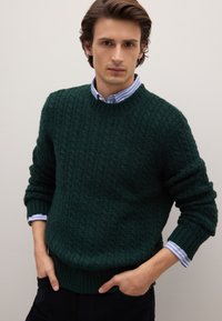 Maglione a coste verde scuro con collo rotondo e polsini a coste, indossato sopra una camicia a righe blu chiaro e bianca.