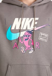 Grå hættetrøje med et stort hvidt "NIKE" logo, farverig grafik af en karakter med solbriller, og et trykt mærke. Medfølgende snorehætte.