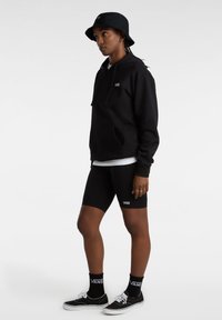Zwarte hoodie met een voorzak, wit logo, in combinatie met zwarte fietsshorts en Vans sneakers. Een zwarte bucket hat maakt de look compleet.