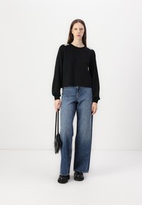 Μαύρη μπλούζα με μακριά μανίκια και διακοσμητικούς ώμους, συνδυασμένη με ψηλόμεσα, φαρδιά τζιν μπλε με λευκές στρας. Το μοντέλο κρατά μια μαύρη τσάντα.