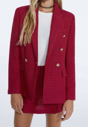 Vrouw draagt een rode, getextureerde, dubbelgeknoopte blazer met gouden knopen over een witte geribde top en bijpassende rode rok.