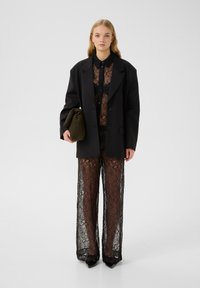Svart överdimensionerad blazer över en genomskinlig spets svart topp och vida byxor. Outfiten inkluderar en grön kuvertväska och spetsiga klackar.