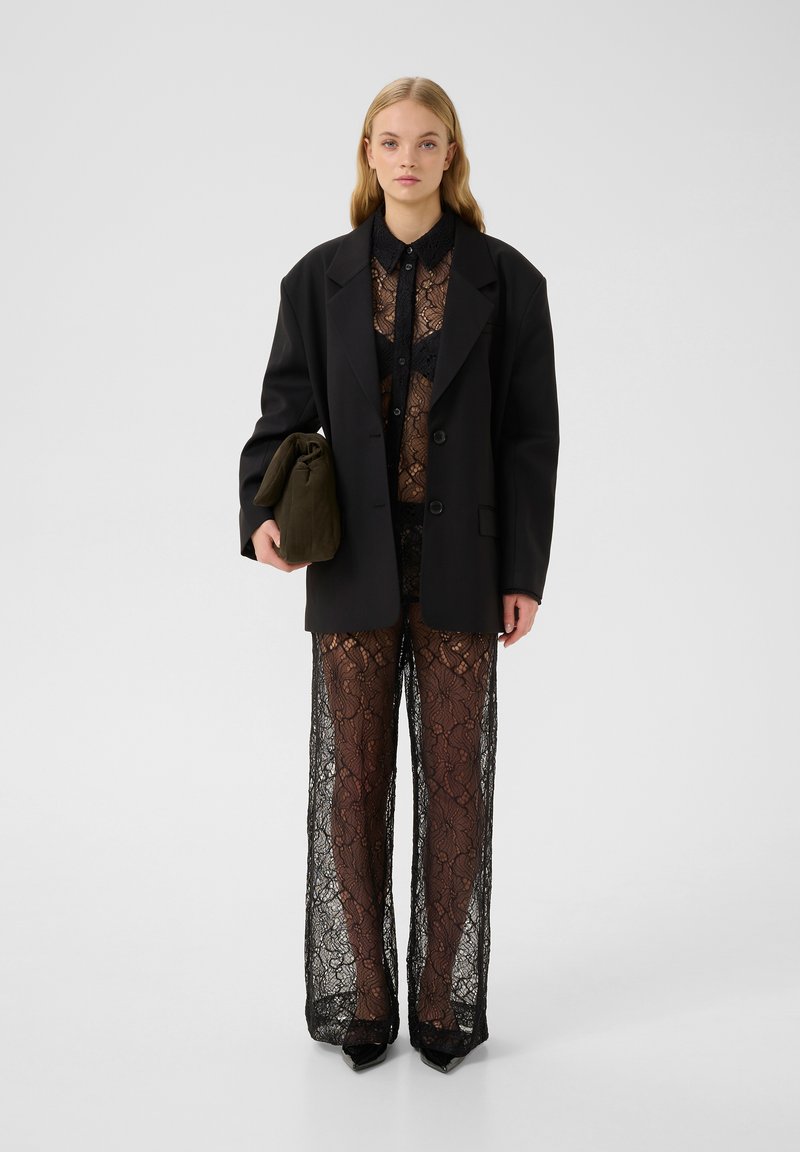 Schwarzer oversized Blazer über einem transparenten, schwarzen Spitzenoberteil und weit geschnittenen Hosen. Das Outfit umfasst eine grüne Clutch und spitze High Heels.