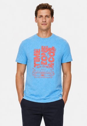 Mann steht frontal und trägt ein blaues T-Shirt mit rotem Grafikdesign, das ein klassisches Auto, Palmen und großen vertikalen Text zeigt.