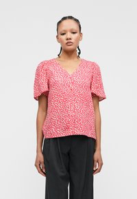 Κόκκινη μπλούζα με λευκές βούλες, V-λαιμόκοψη, κοντά φουσκωτά μανίκια και λεπτομέρεια με κουμπί μπροστά, φτιαγμένη από ελαφρύ ύφασμα. Φορεμένη με σκούρες παντελόνες.