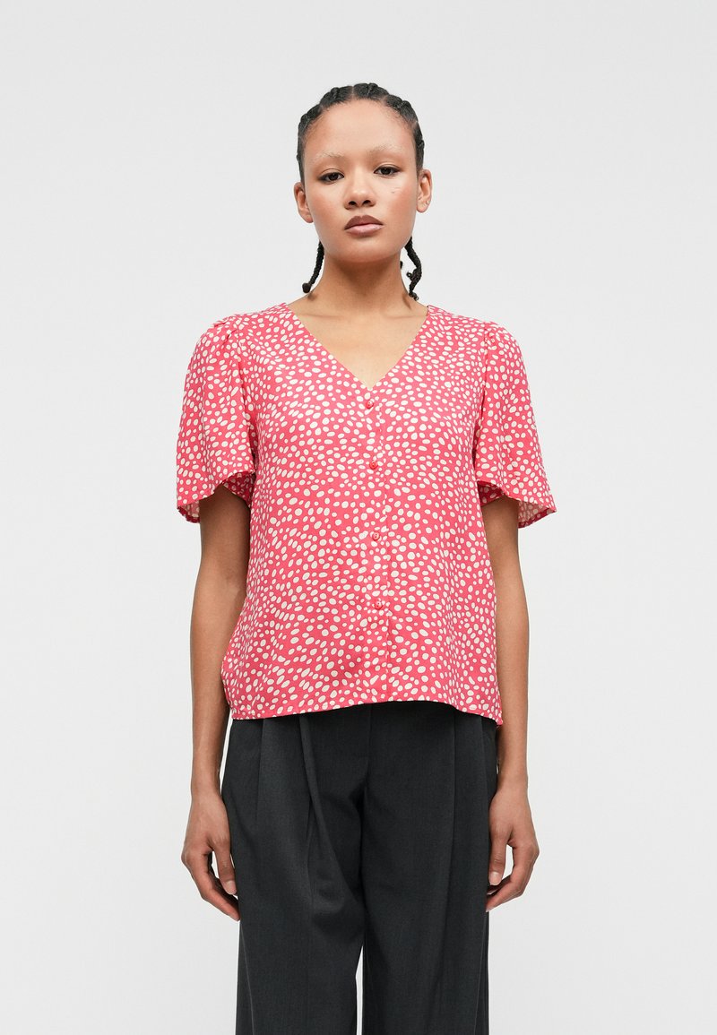 Κόκκινη μπλούζα με λευκές βούλες, V-λαιμόκοψη, κοντά φουσκωτά μανίκια και λεπτομέρεια με κουμπί μπροστά, φτιαγμένη από ελαφρύ ύφασμα. Φορεμένη με σκούρες παντελόνες.