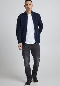 Marineblå ribbet jakke med glidelås over en hvit t-skjorte, parret med mørkegrå jeans og svarte joggesko. Enkelt, moderne og uformelt antrekk.