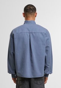Camicia blu a maniche lunghe in morbido tessuto, con colletto rotondo, polsini con bottone e una piega centrale sul retro. Vestibilità rilassata e orlo curvo.