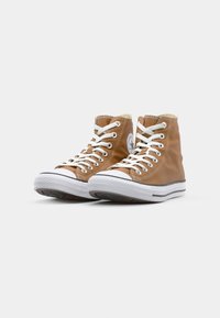 Bruine hoge sneakers hebben witte veters, een rubberen neuskap en een rond logo op de buitenkant. Witte zolen met een gestructureerde loop.
