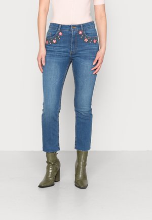 Jeans in denim blu a vita alta con ricami floreali su tasche anteriori, orlo corto e abbinati a stivali con tacco in pelle di coccodrillo verde oliva.