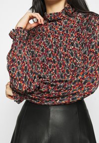 Bedruckte Bluse in dunklem Rot, Grau und Orangetönen mit hohem Kragen. Sie verfügt über geraffte Ärmel und einen lockeren Schnitt, kombiniert mit einem schwarzen Rock.