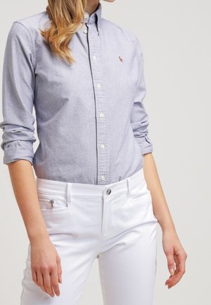 Nainen pukeutuneena vaaleansiniseen napilliseen paitaan, jonka hihat ovat käärittyinä, ja valkoisiin housuihin, joissa on edessä vetoketjullinen tasku. Hän seisoo yksivärisen taustan edessä.