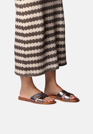 Osoba nosiaca metalické otvorené šľapky a hnedo-béžovú pruhovanú pletenú sukňu, stojaca na svetlom pozadí.