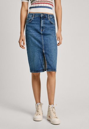 Kvinde iført en blå denim nederdel til knæet med forreste slids, en stribet cropped top og off-white høje sneakers, stående mod en ensfarvet baggrund.