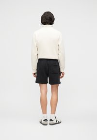 Άτομο με κοντά σκούρα μαλλιά που φορά λευκό μακρυμάνικο κορμάκι, μαύρα σορτς με πίσω τσέπη, λευκές κάλτσες και ασπρόμαυρα αθλητικά παπούτσια, όρθιο, σε όψη από πίσω.