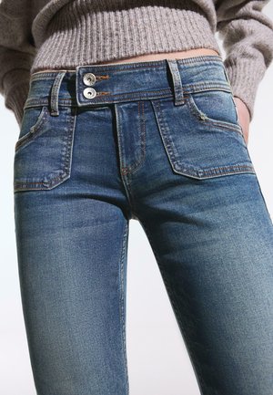 Modré džínsy so stredným pásom, dvojitým zapínaním na gombíky, prednými vreckami a kontrastným šitím, nosené s béžovým rebrovaným svetrom.