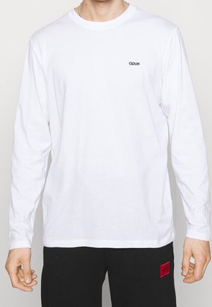 Person wearing a white long-sleeve shirt with small black mirrored logo on chest and black pants with red square patch.