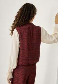 Chaleco con estampado floral burdeos, hecho de una tela texturizada, llevado sobre una camisa de manga larga de color claro con puños rizados. Se muestra la vista trasera.