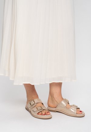 Sandales en daim beige avec deux boucles ajustables et des accents perforés ; portées avec une jupe blanche plissée midi.