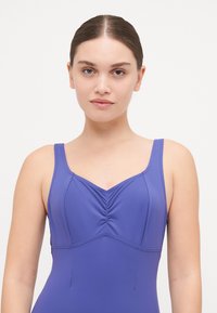 Fialové plavky s priliehavým strihom, ktoré majú široké ramienka, výstrih do srdca a jemné nariasenie na hrudi pre zväčšenie detailu.