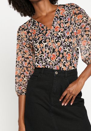 Femme portant un chemisier transparent à fleurs orange et violettes, rentré dans une jupe en jean noire taille haute.