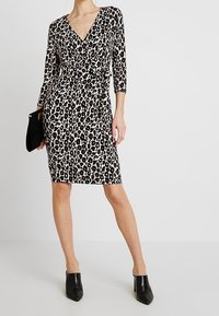 Robe portefeuille imprimé léopard en beige et noir, avec des manches trois-quarts et un décolleté en V, associée à des mules à talons noires et une pochette.