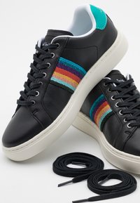 Svarta lädersneakers med flerfärgade randiga textilaccenter, vita sulor och svarta snör. Två svarta skosnören placerade intill.